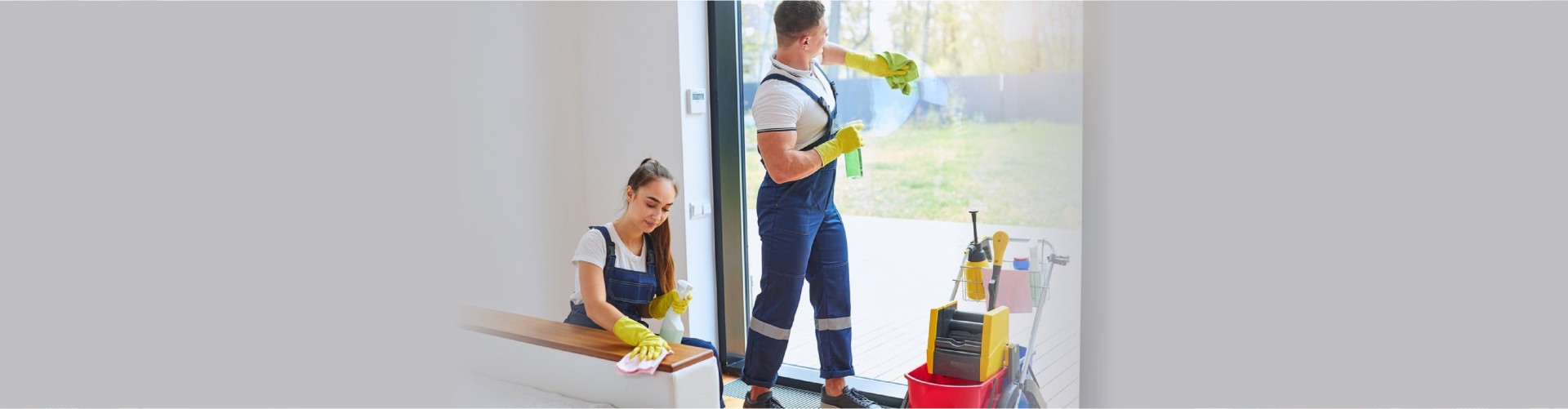 Team of two cleaners effectively cleaning house together, wearing blue uniforms, yellow rubber gloves, men washing window while women wipes dust off sofa